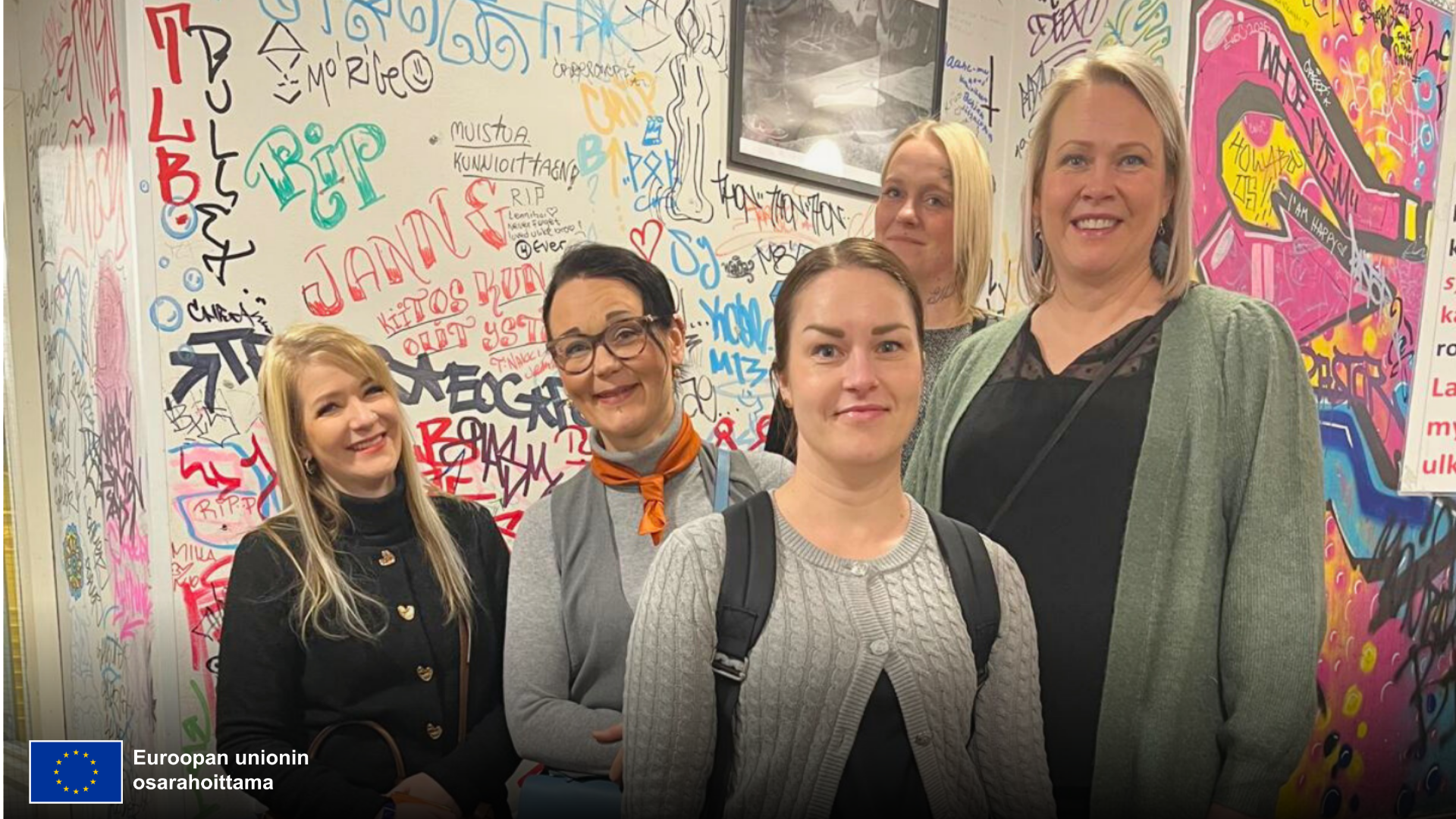 Ryhmä ihmisiä seisoo yhdessä värikkään graffitiseinän edessä. Tausta on täynnä eri väreillä tehtyjä tageja, piirroksia ja tekstejä. Vasemmassa alakulmassa näkyy Euroopan unionin osarahoittamaa -merkintä ja sen vieressä EU-lippu.