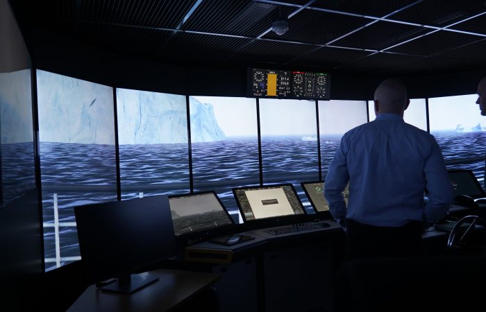 A simulation control room with multiple screens and a wide curved display showing an icy maritime landscape. Two people stand at the control consoles monitoring the simulation.