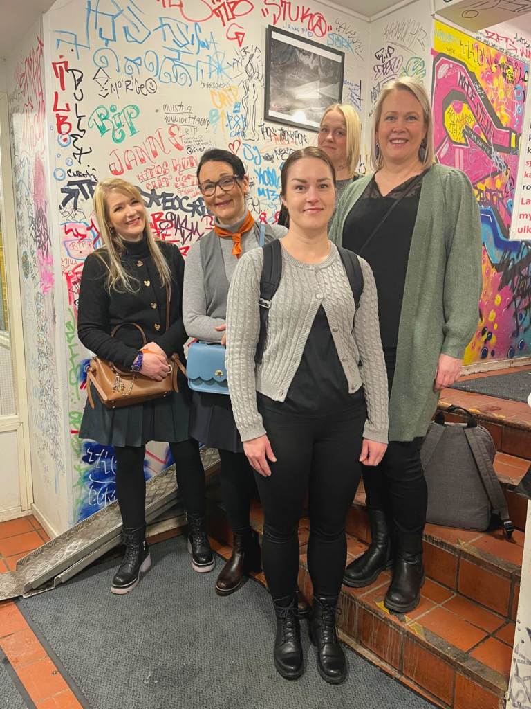 Neljä henkilöä seisoo sisätilassa värikkään graffitiseinän edessä portaikossa.