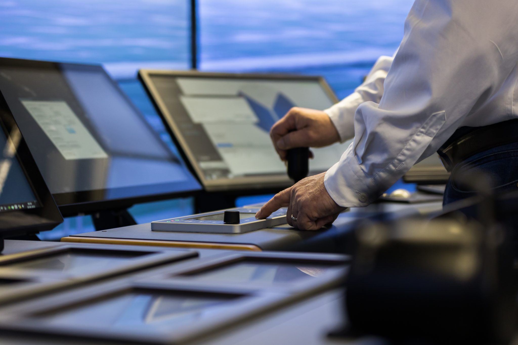 A close-up of a simulator control panel with several touchscreens and an operator’s hand using a control lever. A blue maritime simulation scene is visible on the display in the background.