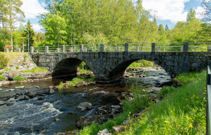 Valokuva vanhasta kivisestä kaarisillasta, joka ylittää virtaavan joen metsäisessä ympäristössä.