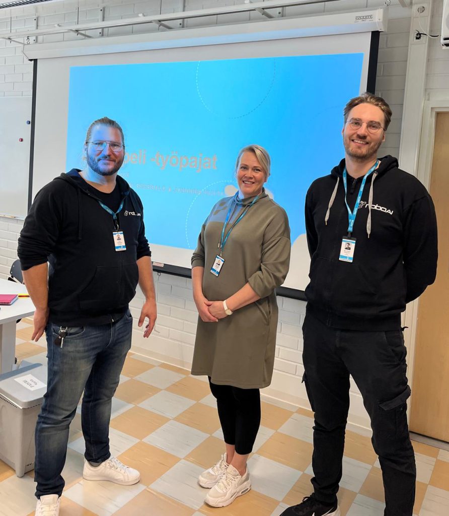 Kolme henkilöä seisoo luokkatilassa esitysnäytön edessä.