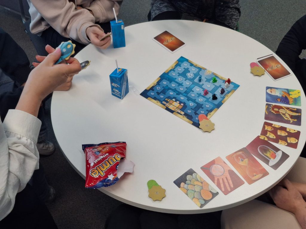 Pöydän äärellä pelataan lautapeliä, pöydällä on kortteja ja välipaloja.