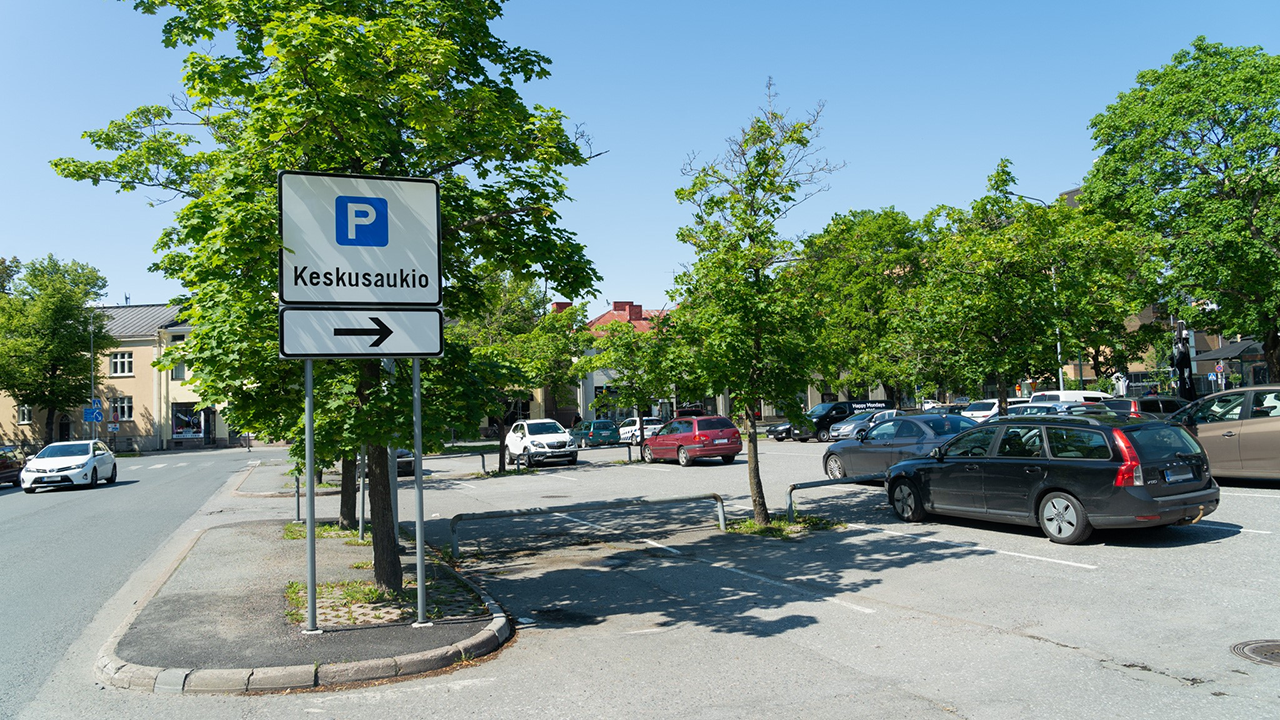 Parking area with cars and a road sign pointing to ‘Keskusaukio,’ surrounded by trees and buildings on a sunny day.