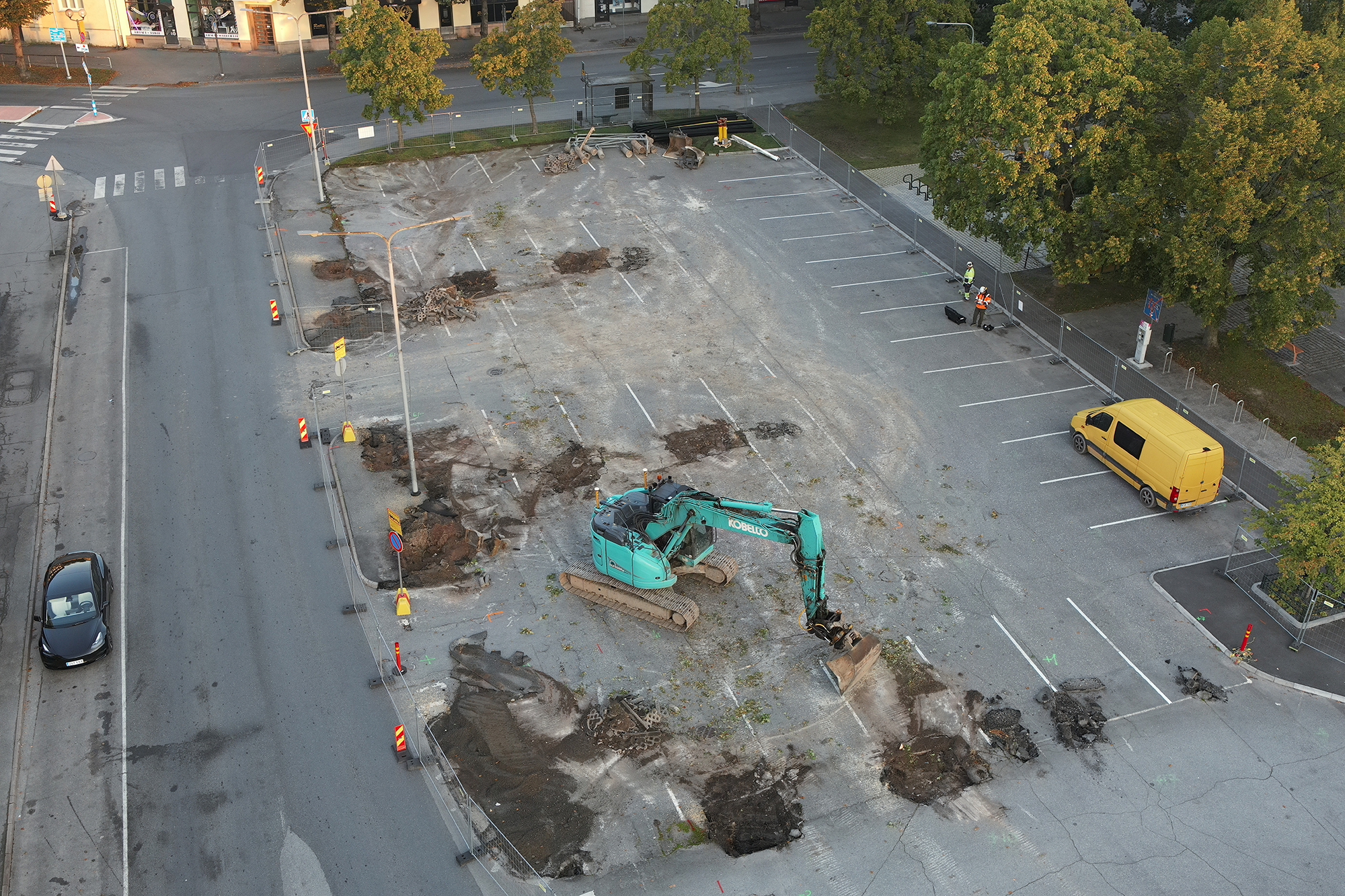 Aerial view of a fenced city parking lot under construction, with an excavator removing asphalt and workers on site, surrounded by trees and streets.