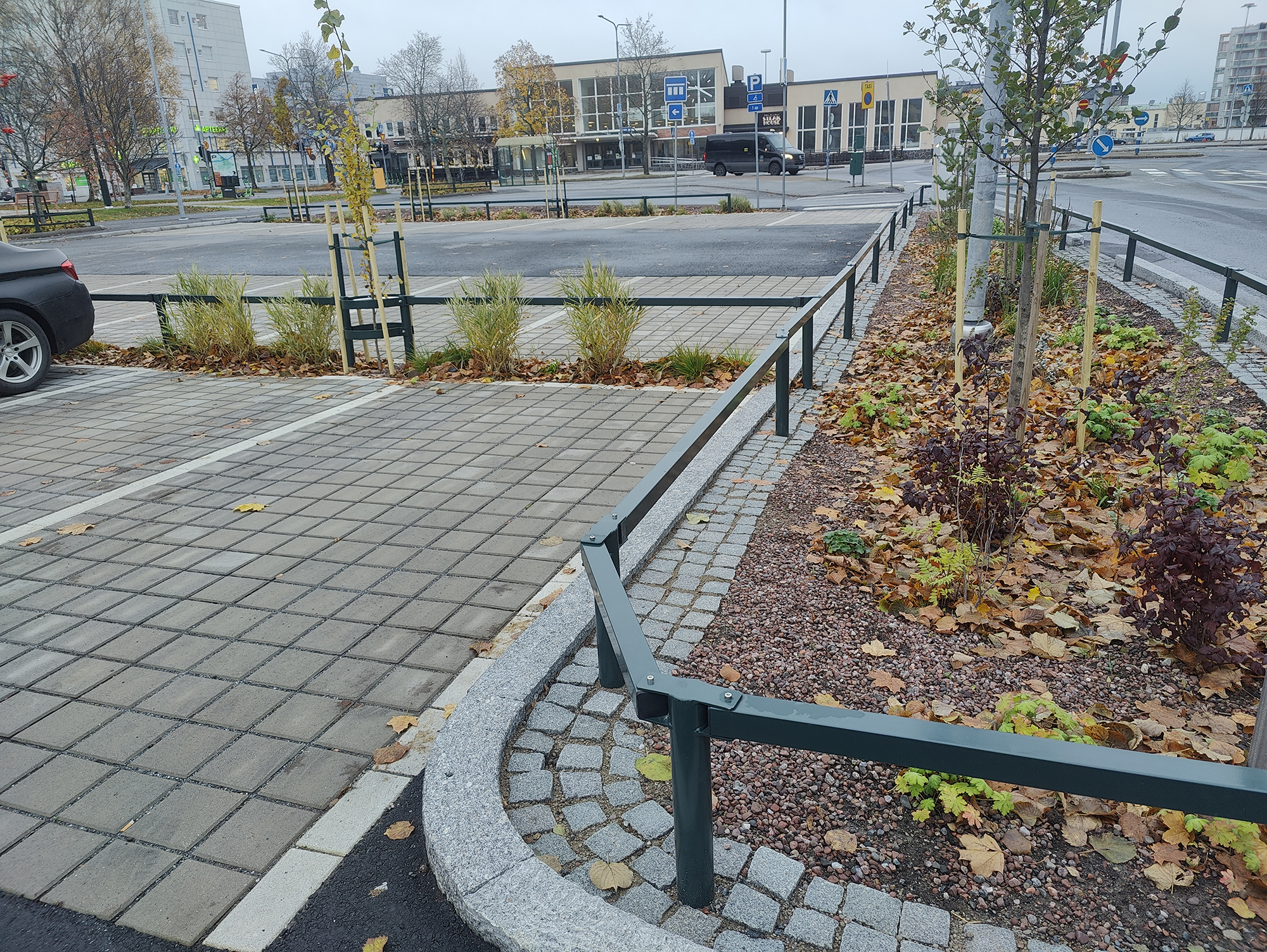 Urban pedestrian area with paved walkways, small planted trees, and low metal railings near a street intersection on an overcast autumn day.