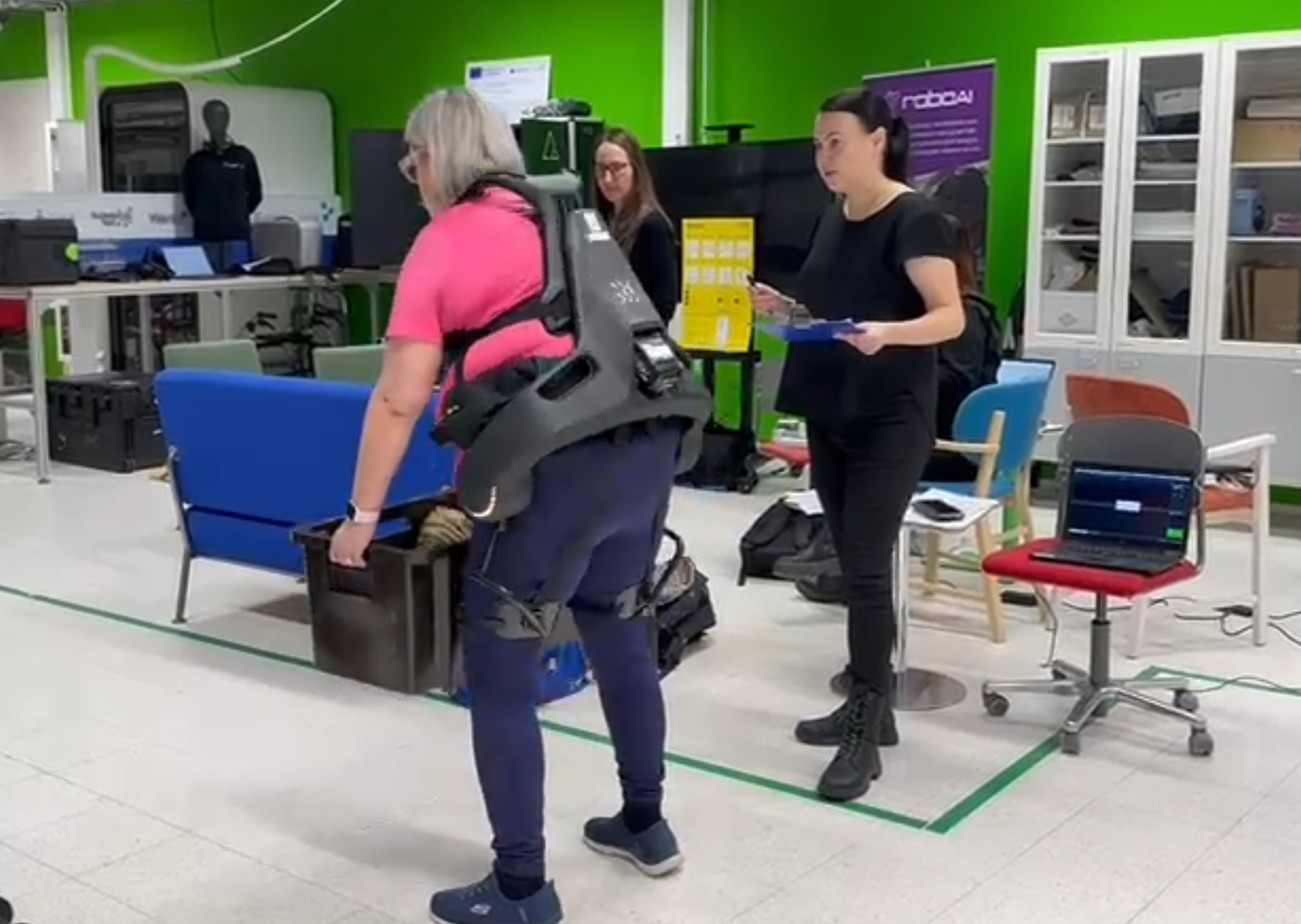 Henkilö käyttää eksoskeletonia ja nostaa laatikkoa laboratoriotilassa, vieressä tutkija seuraa tilannetta ja mittauslaitteisto on valmiina.