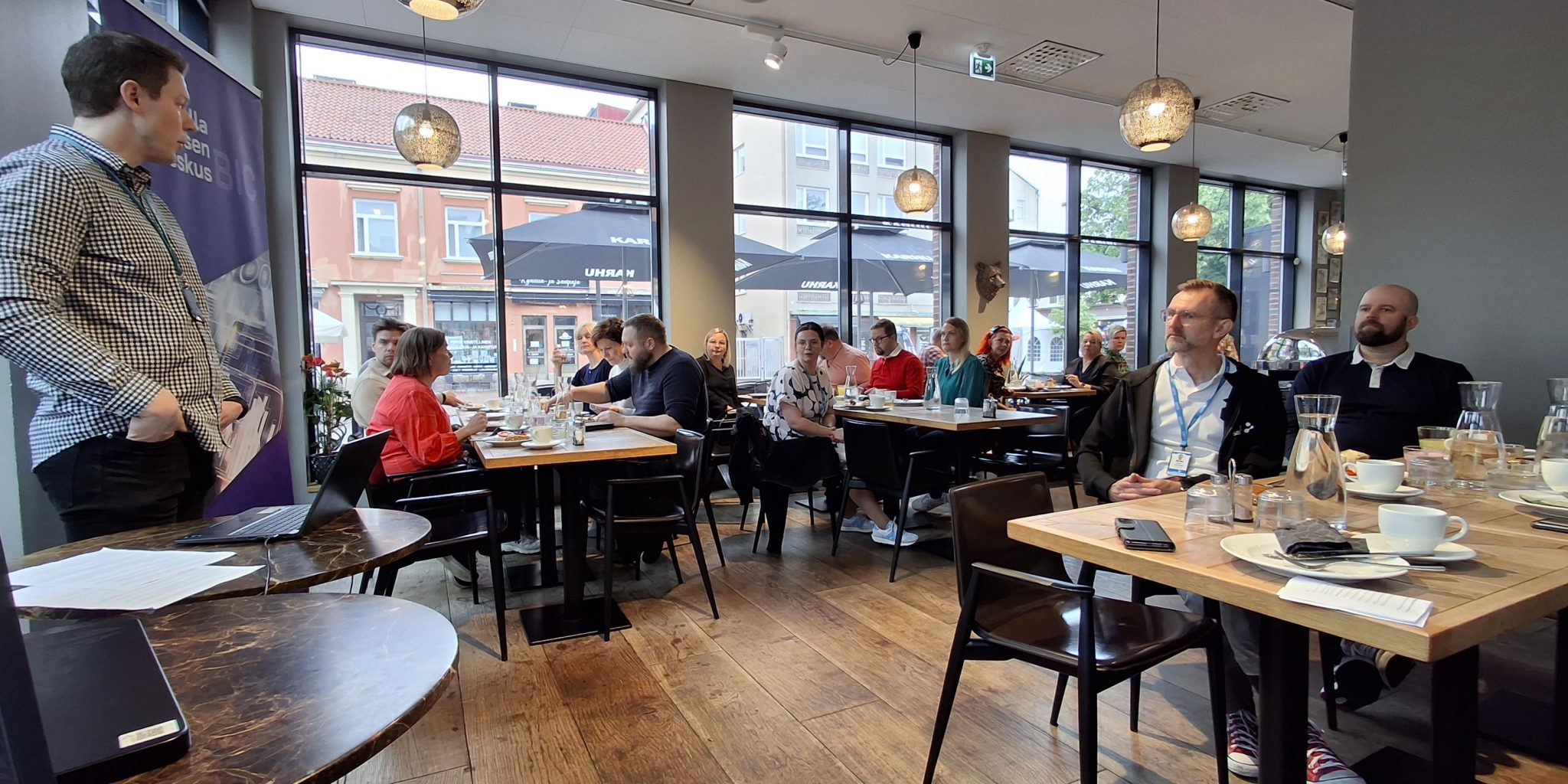 yleiskuvaa brunssitilaisuudesta. Ihmisiä istumassa pöytien ääressä ja nauttimassa brunssin antimista.