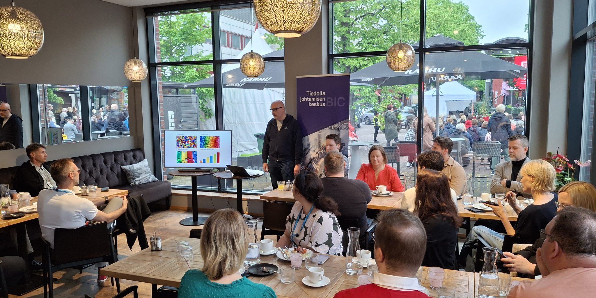 Jani Nurmi pitää esitystä. Ravintolan sisätiloissa pöytien ääressä istuu ihmisiä kuuntelemassa.