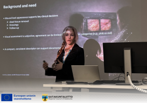 Mitra Daneshmand pitää esitystä luokkatilassa ja seisoo tietokoneen sekä näytön vieressä. Taustalla on dia, jossa käsitellään haavan arviointia ja kliinisiä päätöksiä, kuten kuolleen kudoksen poistamista, sidoksia ja seurantaa. Alareunassa näkyvät Euroopan unionin ja Satakuntaliiton logot.