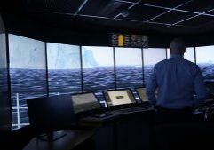 A simulation control room with multiple screens and a wide curved display showing an icy maritime landscape. Two people stand at the control consoles monitoring the simulation.