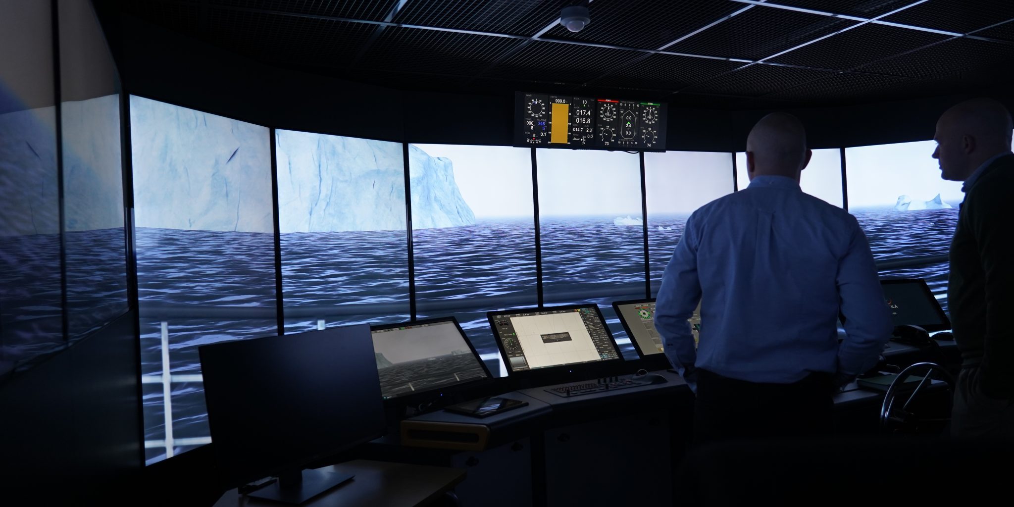 A simulation control room with multiple screens and a wide curved display showing an icy maritime landscape. Two people stand at the control consoles monitoring the simulation.