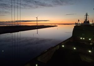 Rahtialus satamassa auringonlaskun aikaan, taustalla tuulivoimala.