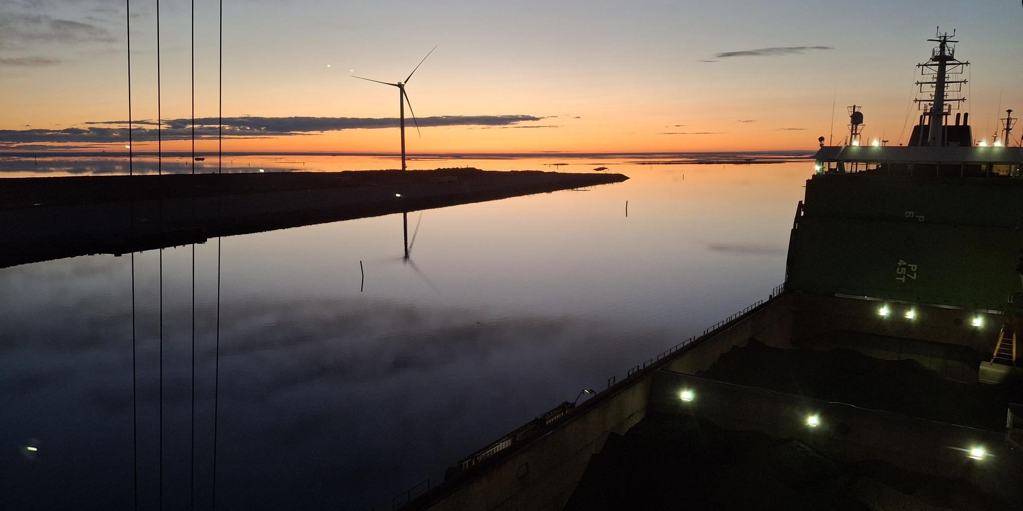 Rahtialus satamassa auringonlaskun aikaan, taustalla tuulivoimala.