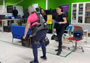 Henkilö käyttää eksoskeletonia ja nostaa laatikkoa laboratoriotilassa, vieressä tutkija seuraa tilannetta ja mittauslaitteisto on valmiina.