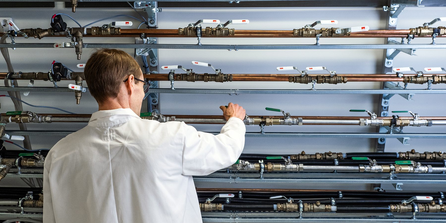 Laboratoriotilanne, jossa henkilö valkotakissa tarkastelee seinälle asennettuja putkirivejä ja venttiilejä.