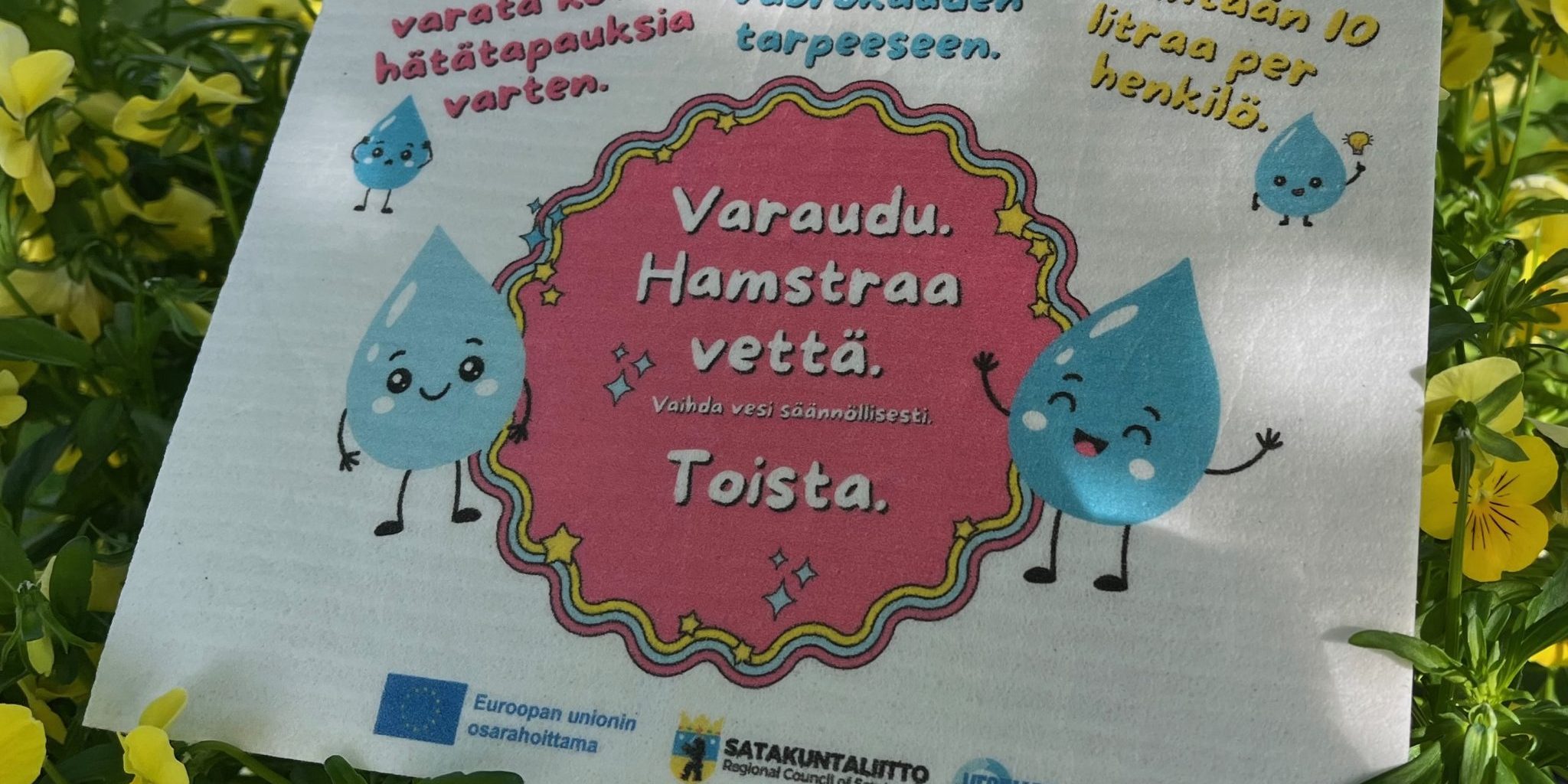 Vesivara-tiskirätti kukkien keskellä muistuttaa kotitalouksien vesivarautumisesta häiriötilanteissa.
