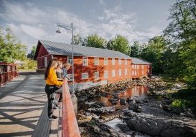 Ihmisryhmä seisoo puusillalla katsomassa alapuolella virtaavaa kivikkoista jokea. Taustalla on suuri punainen puinen rakennus ja ympärillä vihreää puustoa.