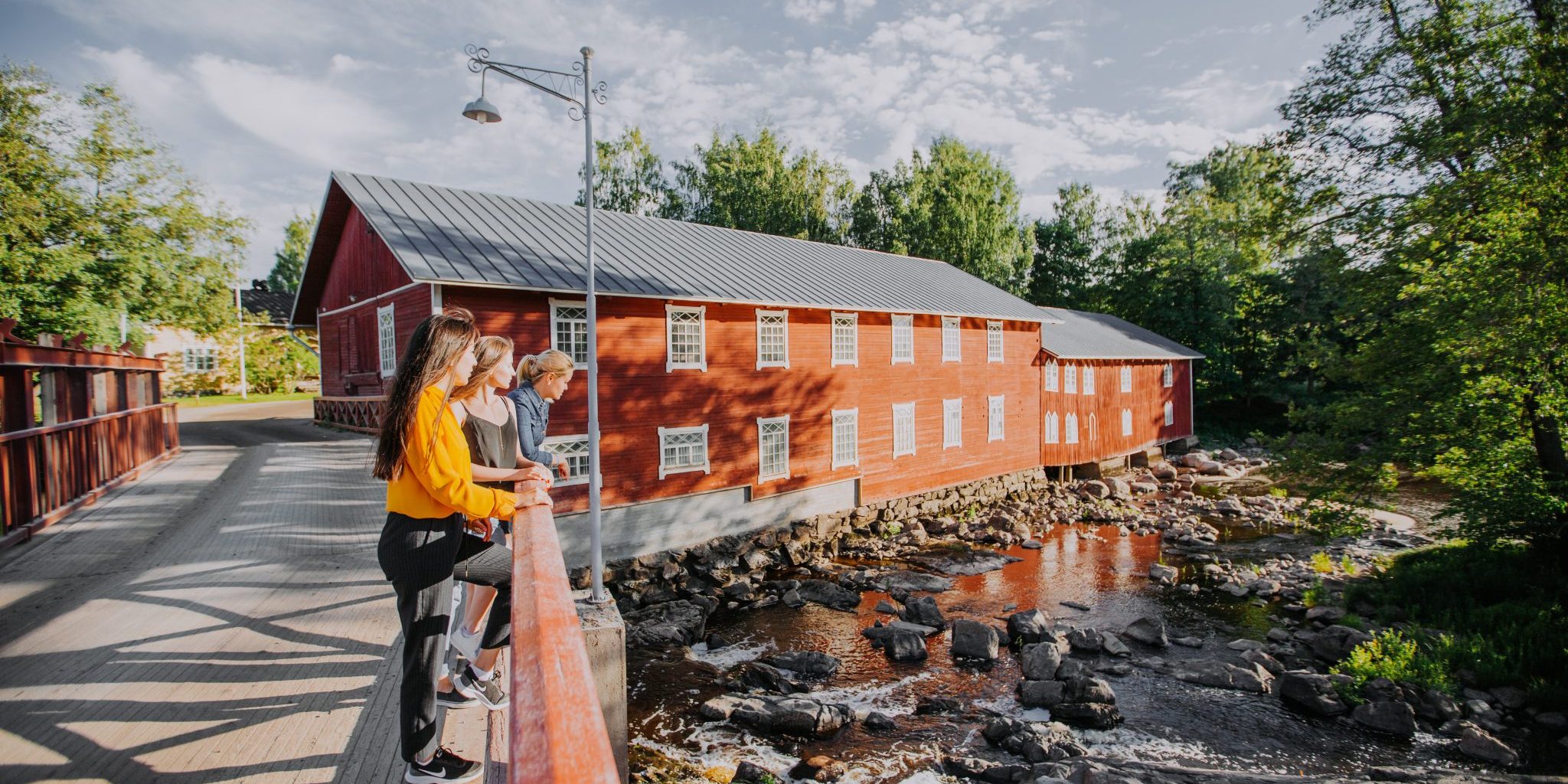 Ihmisryhmä seisoo puusillalla katsomassa alapuolella virtaavaa kivikkoista jokea. Taustalla on suuri punainen puinen rakennus ja ympärillä vihreää puustoa.