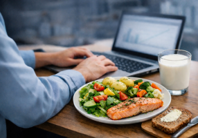 Lautasellinen ruokaa, jossa on lohta, perunoita ja salaattia, tietokoneen ääressä työpöydällä. Vieressä lasi maitoa ja voideltu leipä.