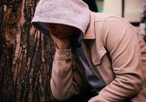 Huppariin ja takkiin pukeutunut henkilö istuu puun juurella käsi poskeaan vasten.