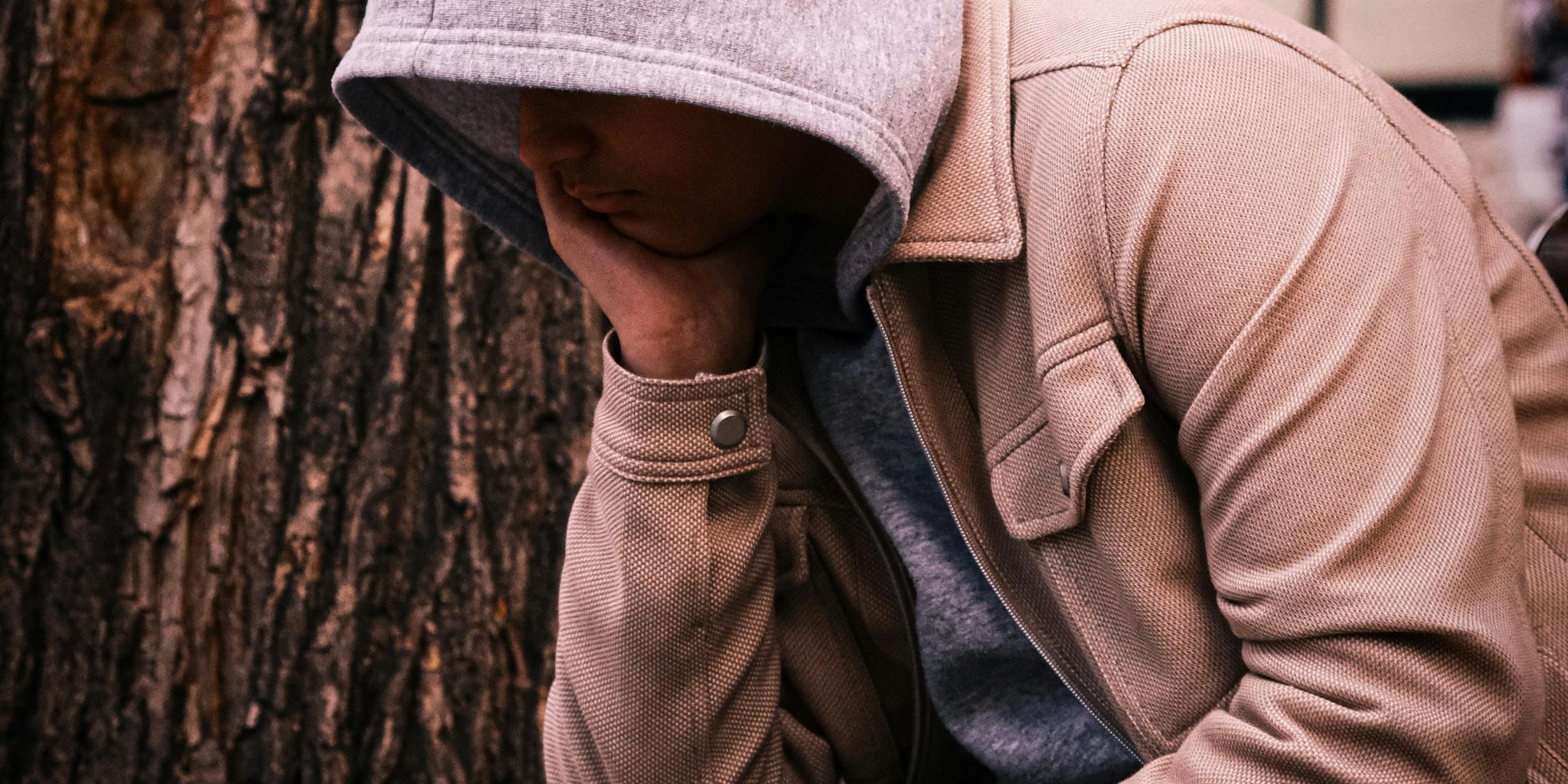 Huppariin ja takkiin pukeutunut henkilö istuu puun juurella käsi poskeaan vasten.