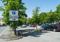 Parking area with cars and a road sign pointing to ‘Keskusaukio,’ surrounded by trees and buildings on a sunny day.