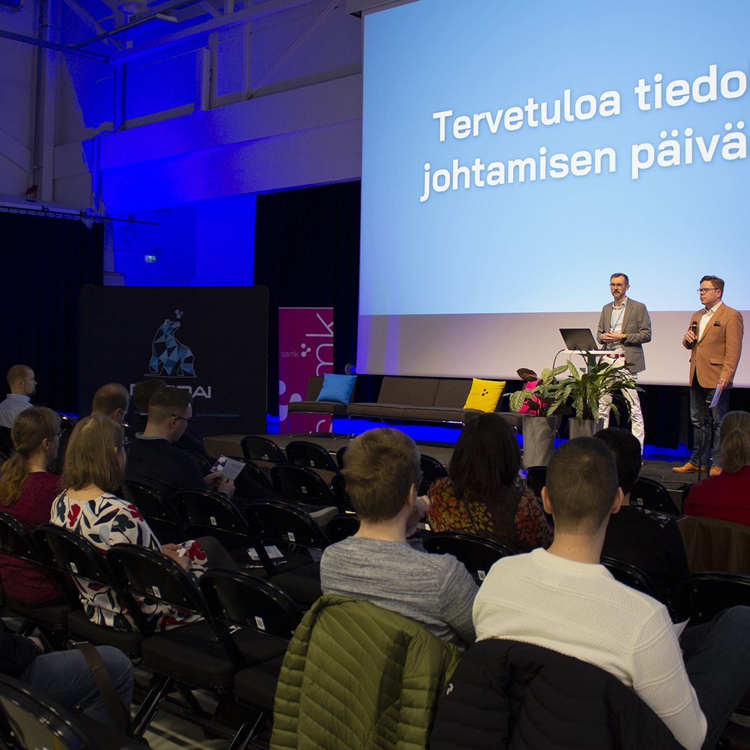 Kuva tiedolla johtamisen tapahtumasta, lavalla kaksi henkilöä, joita yleisö seuraa.