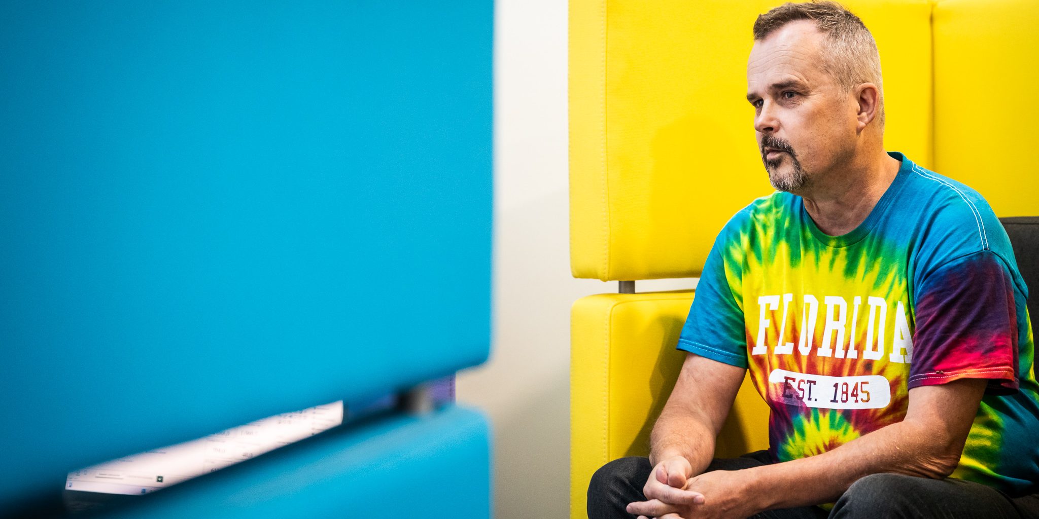 Kuvassa oikealla mies värikkäässä paidassa istumassa mietiskelevän näköisenä.