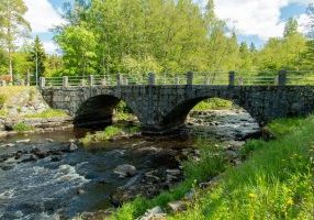 Valokuva vanhasta kivisestä kaarisillasta, joka ylittää virtaavan joen metsäisessä ympäristössä.