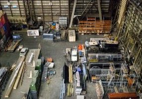 Teollinen varastohalli, jossa on järjestettyinä metallirakenteita, rakennusmateriaaleja ja ajoneuvoja suurina pinoina lattialla.